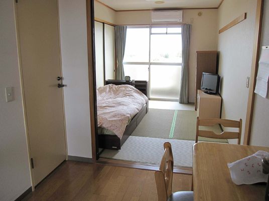 テレビや寝具付きのベッドが置かれた部屋にはエアコンも付き、手前のフローリングの部屋にはテーブルセットが置かれている。