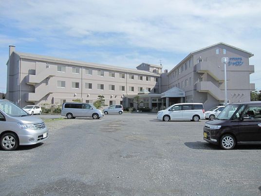 エル字型の3階建ての建物はベージュの外観で、敷地内には駐車場があり、車が数台止められている。
