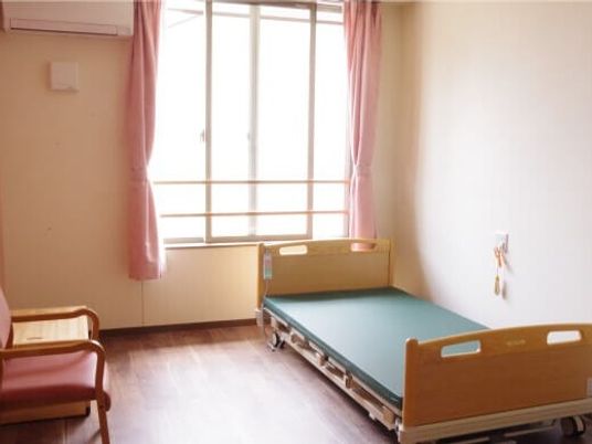 緊急通報装置がベッドサイドにあり、介護ベッドやエアコンが付いたフローリングの部屋。