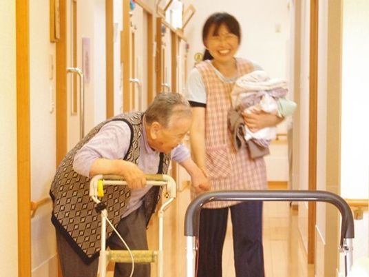 手をつないで移動している施設スタッフと歩行器具を利用している高齢者の様子。笑顔で会話している
