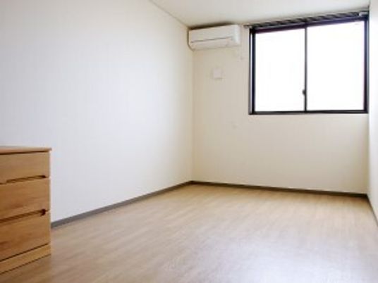 フローリングの部屋にはカーテンのついていない大きな窓があり、日当たり良好。壁際には木製のタンスが置かれ、壁にはエアコンが付いている。