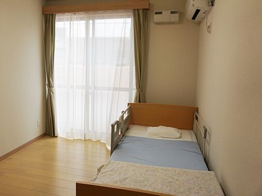 レースカーテンとカーテンが付いた大きな窓が付いているフローリングの部屋には寝具付きのベッドが置かれ、エアコンもある。