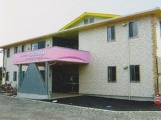 ホーム外観。２階建ての建物を斜め左前から撮影。壁の色は淡いベージュ、屋根は三角形で黄色、玄関の上にはピンクの雨よけ用の屋根がある。