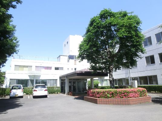白を基調とした３階建ての建物の前と周りには草木が植えられ、玄関には車寄せが付いている。