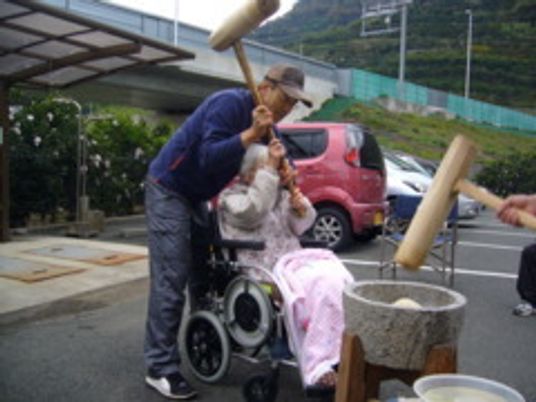 車いすの女性入居者の手を取り、一緒に餅つきを楽しむ男性。