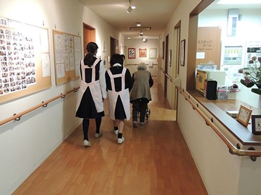 廊下を歩く高齢者とスタッフ廃りの後ろ姿。施設内の廊下の一場面