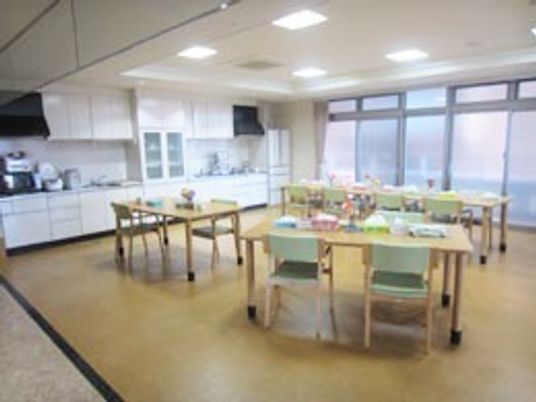 大きなキッチンが付いた食堂は一面が窓で明るい。家庭サイズのテーブルが複数並び、グリーンのいすがセットされている。