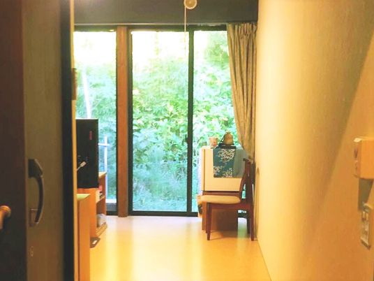 緑あふれる風景が大きな窓の外に広がっている。カーテンは開けられ明るい雰囲気の居室内には、いすなどの私物が持ち込まれている。