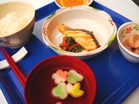 麩が浮いた吸い物や白いご飯、煮物に焼き魚、カットフルーツなどが並んだブルーのトレー。