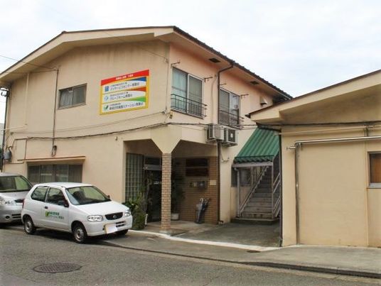 ２階建ての建物の前には車が２台止まり、横には屋外階段がある。