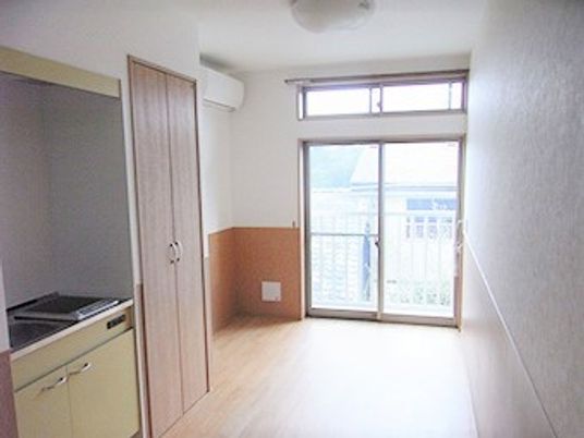 フローリングの部屋。大きな窓にはカーテンが付き、キッチンやエアコン、収納家具もある。