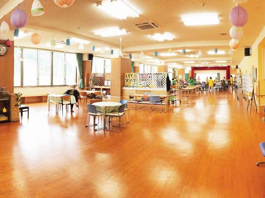 広々としたホールにはたくさんの提灯が飾られ、大きな窓がある。複数のダイニングテーブルセットがあり、入居者が1名椅子に座っている。