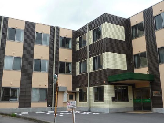 3階建てのエル字型の建物はベージュと黒で玄関には屋根があり、敷地内には駐車場がある。