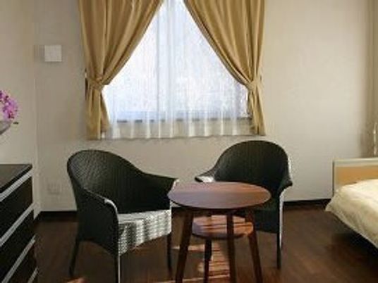 テーブルセットやベッドが置かれたフローリングの部屋には、レースカーテンとカーテンが付いた窓がある。