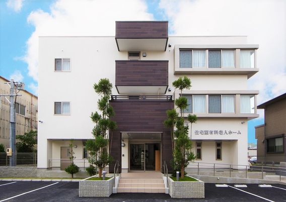 白を基調とした明るい3階建ての建物。各フロアにブラウンのベランダが付き、玄関には手すり付きのスロープがあり、駐車場もある。