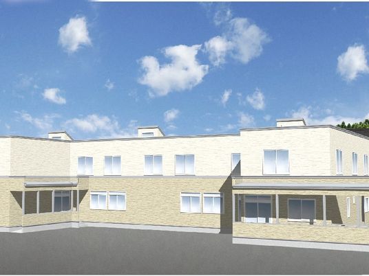 2階建ての建物は横に長く、1階部分はベージュ、2階部分は白で周辺には緑も多い。