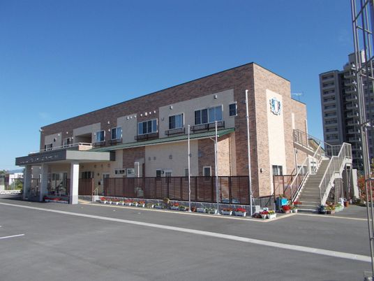 ベージュとブラウンの2階建ての建物。建物の周りには赤い花が植えられたプランターが並べられ、屋外階段や車寄せも付いている。