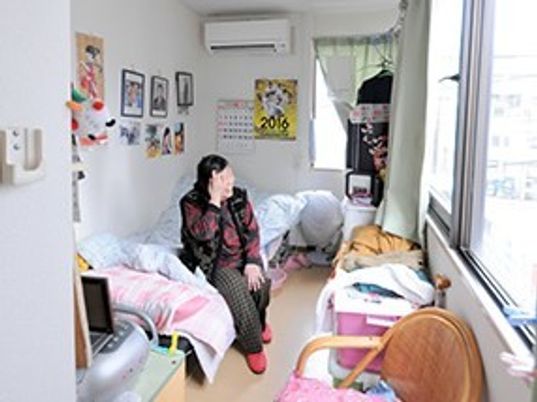 女性入居者がベッドに座り、居室には椅子やエアコン、個人の写真などが置かれている。