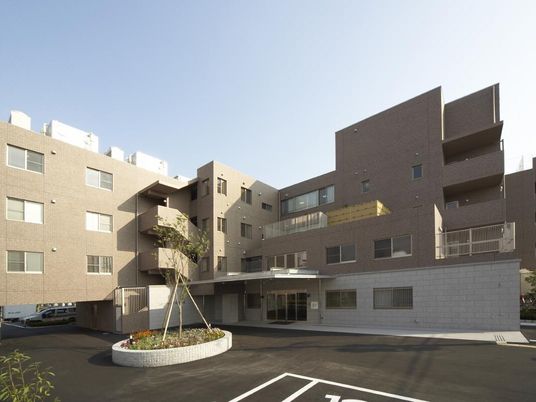 ブラウン系の４階建ての建物の前には駐車場があり、草木が植えられている。