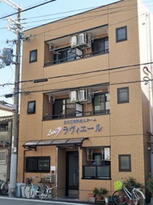 ブラウン系の建物は3階建て。自転車などが置かれた入り口の上部の外壁にはホーム名が書かれている。