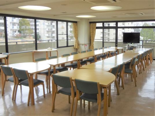 長机と椅子が複数並べられている施設内のリビング。テレビが奥にある様子