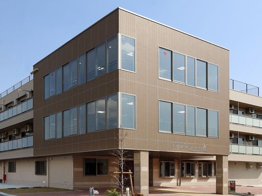 ３階建ての建物はブラウン系で各フロアにベランダがあり、建物の前には木が植えられている。
