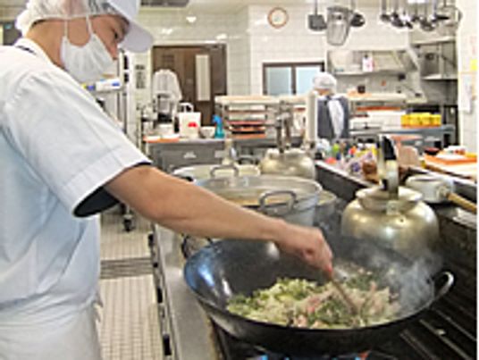 大きな中華鍋でいためものを作る調理スタッフ。白いマスクやネット付きの帽子をしている。