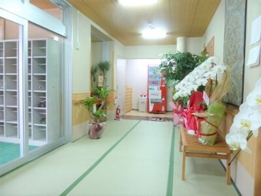 畳が敷かれたロビーにはお祝いの花や観葉植物、自動販売機などが置かれ、自動ドアの外には靴箱もある。