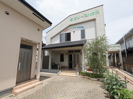 白い外壁の2階建ての建物。外壁には住宅名が書かれ、玄関からは手すり付きの階段があり、菜園スペースもある。
