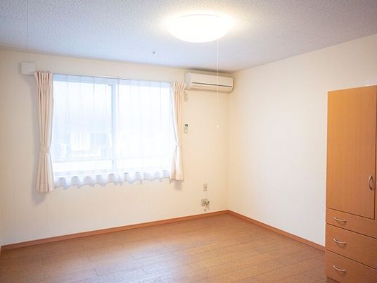 エアコンや大型の収納家具が付いたフローリングの部屋にはレースカーテンとカーテンが付いた大きな窓がある。