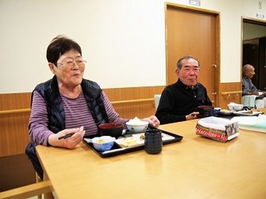 眼鏡をかけた男女の入居者が楽しそうに食事をしている様子。木製のテーブルにはトレーに乗った食事が並んでいる。