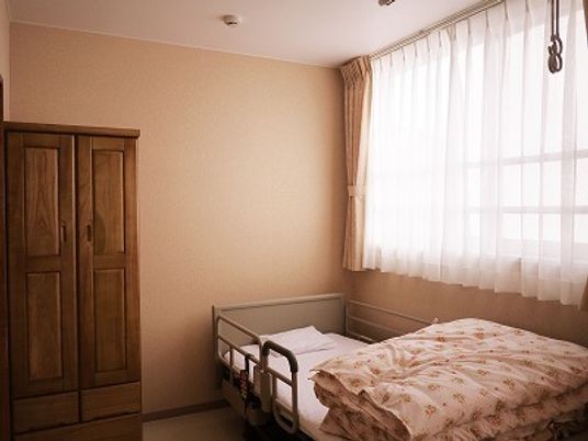 寝具付きのベッドが置かれたフローリングの居室には家具もあり、大きな窓が付き明るい雰囲気。