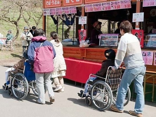 スタッフの介助を受けながら外出を楽しむ車いすの入居者2名。屋台が並ぶ公園で。