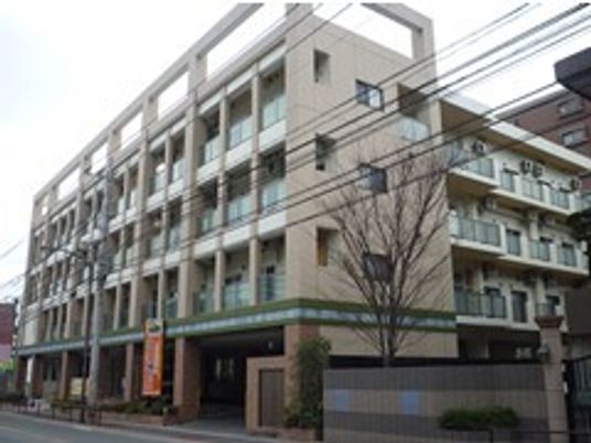 ベージュの外観の４階建ての建物の周りには草木が植えられている。各フロアにベランダがあり、重厚感のある造り。