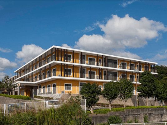 オレンジの外壁の3階建ての建物。玄関先には車寄せが付き、周りには草木が植えられている。