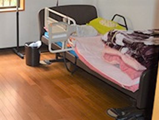 フローリングの洋室の居室。手すり付きのベッドには布団や毛布が積み上げられている。