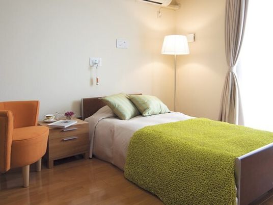 寝具がセットされたベッドや一人用の椅子が置かれたフローリングの部屋には、背の高いライトがあり、エアコンや緊急通報装置もある。