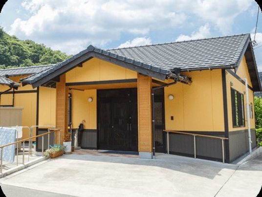 オレンジの外壁の平屋建ての建物。玄関の周りには手すりがあり、玄関には屋根があり、周辺には緑が多くある。