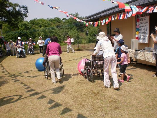 敷地内で運動会を楽しむ入居者とスタッフ。ボールを追って車いすで動く入居者をサポートするスタッフ。
