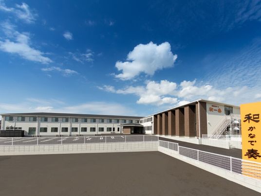 青空の下に建っている２階建ての建物はエル字型で広い駐車場が付いている。外壁にはホーム名が書かれている。