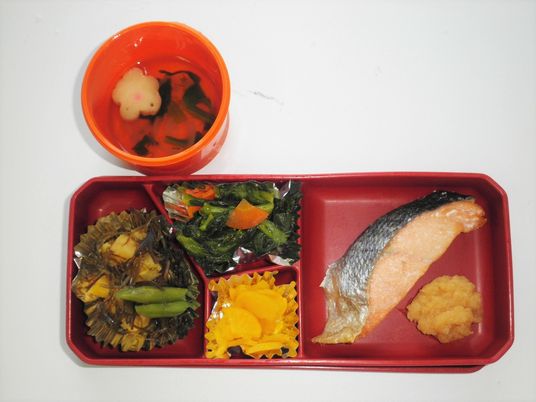 焼き鮭や野菜のあえ物、漬物などが詰められた弁当風の食事に椀が添えられている。