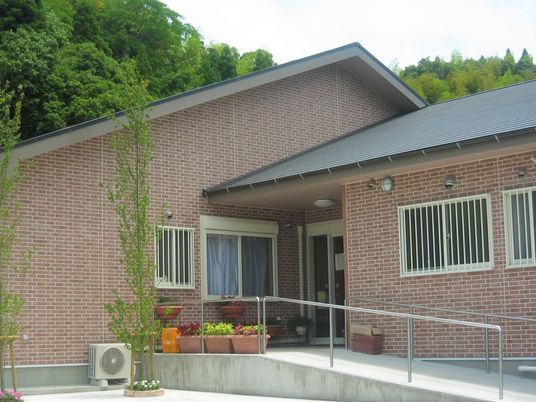 ブラウンのレンガ調の外壁の平屋建ての建物は明るい雰囲気で、玄関先には花が植えられたプランターが置かれ、駐車場もある。