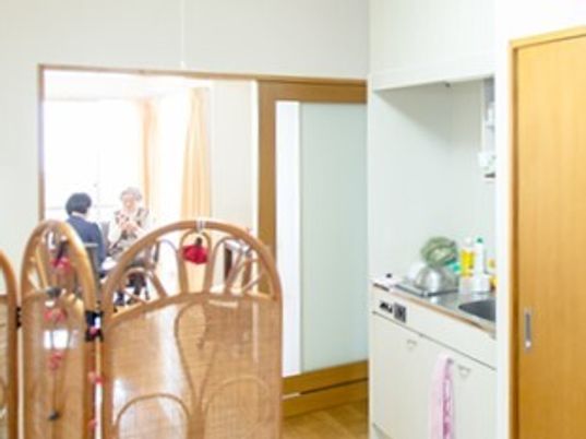 手前には調理用具が置かれたキッチンがあり、タオルがかかっている。奥のリビングスペースでは女性が2名ティータイムを過ごしている。