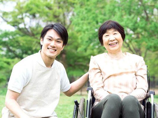 自然が広がる場所で車いすに乗りリラックスする女性とスタッフ。カメラに笑顔を向けている様子