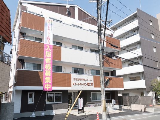白とブラウンの4階建ての建物にはベランダがつき、玄関には手すり付きのスロープがあり、周りにも背の高い建物がある。