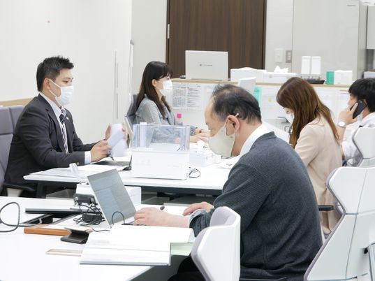 向かい合わせにデスクをくっつけて仕事に当たっている男女のスタッフ陣。スタッフの年齢はバラバラで、それぞれにパソコンがあり、マスクを付けている。