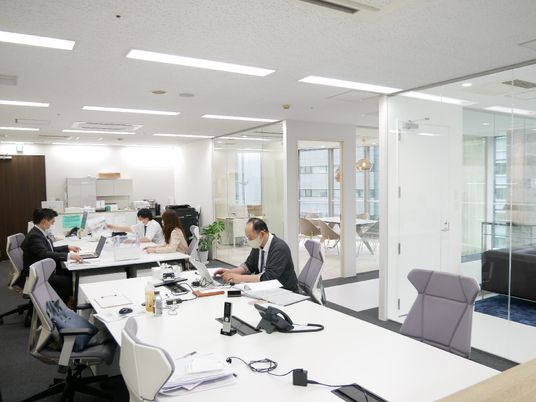 白いデスクが向かい合わせに設置されている。それぞれのデスクにはグレーの椅子がセットされ、電話やパソコンが置かれている。