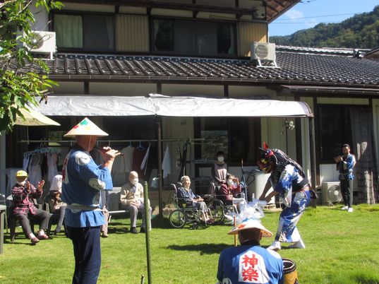 芝生が敷かれた庭で獅子舞を見ている入居者たち。青いハッピを着た男性たちが笛を吹いたり太鼓をたたいたりしている。