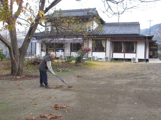 広い敷地内には大きな木が植えられ、落ち葉が落ちている。それを高齢男性が一人で集めている様子。奥には和風の民家がある。