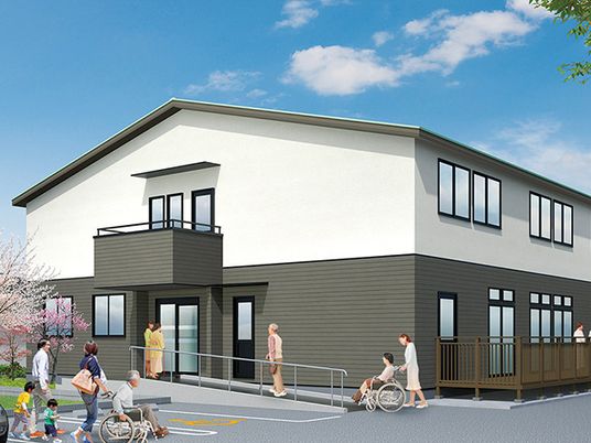 モノトーンの2階建ての建物。玄関には手すり付きのスロープがあり、共有スペースから出入り可能なウッドデッキのテラスもある。